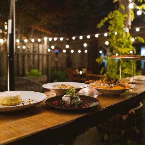 Plates of food line a long table on an outdoor patio. String lights hang over the patio.
