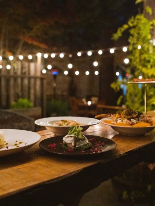 Plates of food line a long table on an outdoor patio. String lights hang over the patio.
