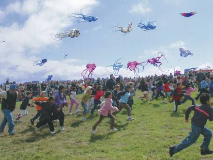 Berkeley Kite Festival attendees, mostly children, run in the grass beneath blue skies as multicolored kites fly overhead.