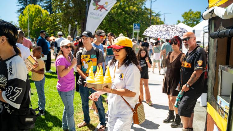 A group of people stand in the sun, grabbing food from a food truck that's been parked on bright green grass in Berkeley, CA