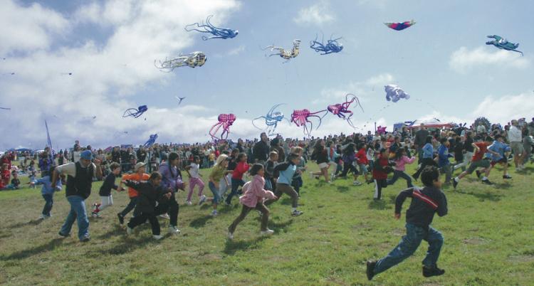 Berkeley Kite Festival attendees, mostly children, run in the grass beneath blue skies as multicolored kites fly overhead.