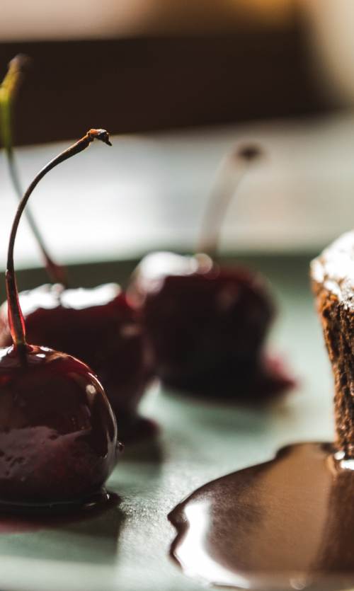 A chocolate cake dessert, topped with a scoop of ice cream and chocolate syrup, sits on a plate next to cherries.