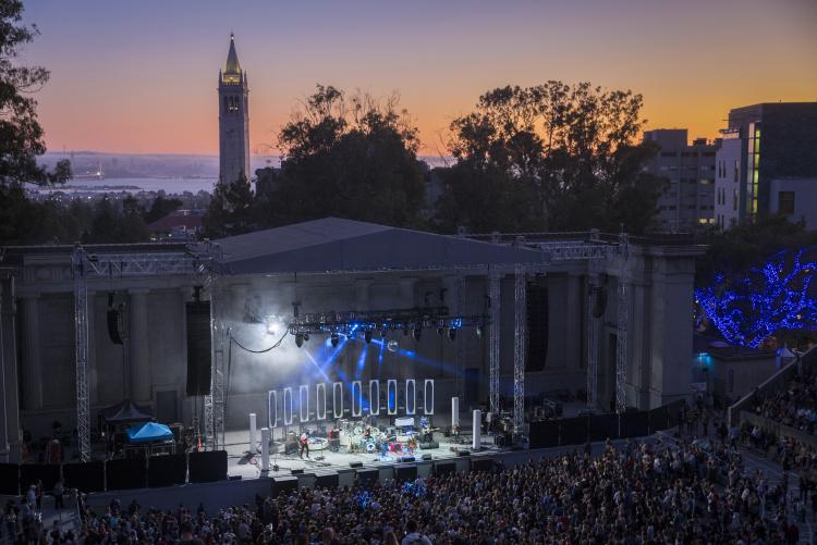 A large crowd watches a band perform on the Greek Theatre stage. The Berkeley skyline is visible behind the stage