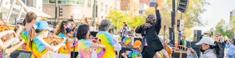 The conductor and musicians of Berkeley Symphony wear tie dye shirts while performing outdoors for a crowd