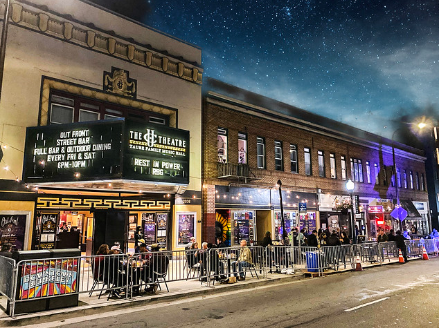 People sit at tables outside of the UC Theatre at night. The theatre has a black marquee and multiple show posters