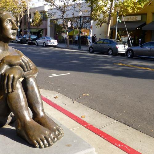 Bronze statue of a person sitting at Fourth Street in Berkeley, CA. Shops are visible across the street